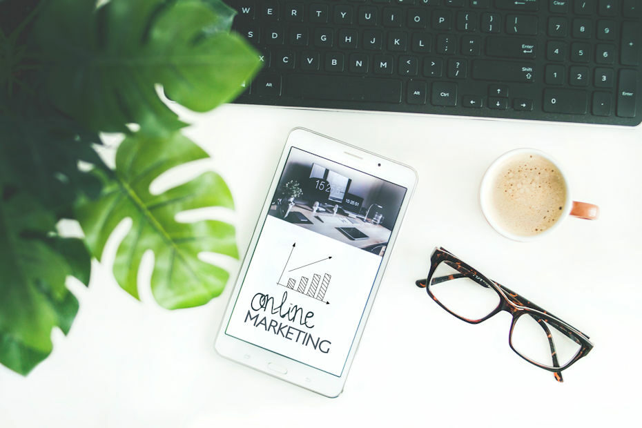 On a white table sits a keyboard, a cup of coffee, a pair of eyeglasses, a green leafy plant, and a tablet. The tablet shows a page with the words "online marketing" as a way for monetizing your content.