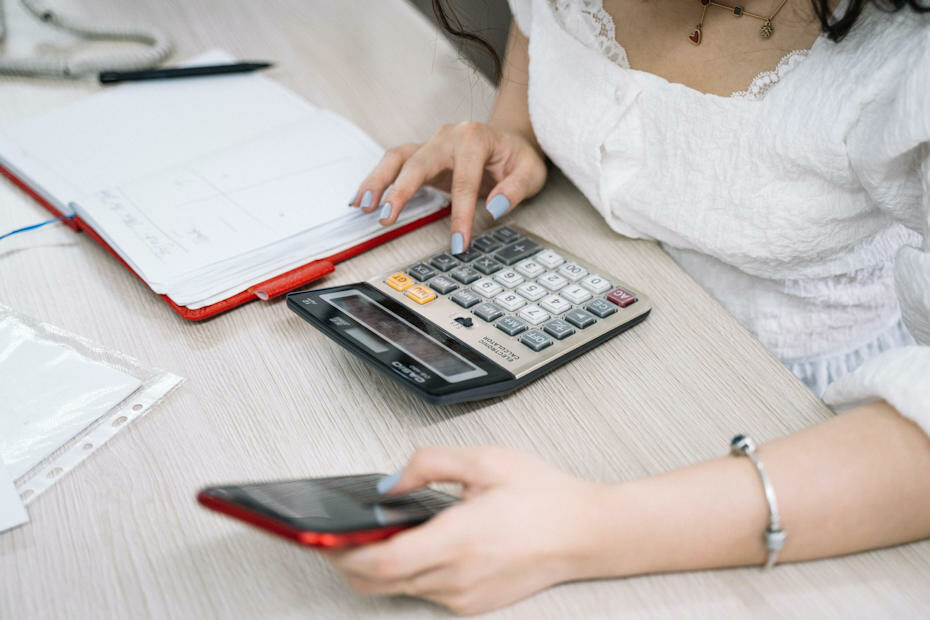 A woman sits at a table holding a red smartphone in her left hand while punching numbers on a calculator with her left hand. A red notebook lies open close to her right hand. She is figuring out her influencer rate before giving a quote to an advertiser.