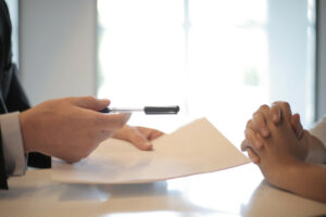 A closeup photo of a man's hands to the left of the photo showing him holding a pen out with his right hand and pushing some paperwork with his left hand towards the hands of a woman on the right side of the photo. The woman's hands are clasped and she's not accepting the paper or the pen. She's not taking the first offer and will stay tough as she negotiates her influencer contract.