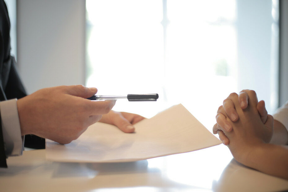 A closeup photo of a man's hands to the left of the photo showing him holding a pen out with his right hand and pushing some paperwork with his left hand towards the hands of a woman on the right side of the photo. The woman's hands are clasped and she's not accepting the paper or the pen. She's not taking the first offer and will stay tough as she negotiates her influencer contract.