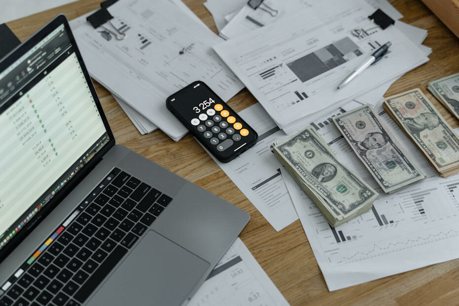 A desk with a laptop to the left, a calculator at the center, 4 piles of cash neatly stacked, and piles of paper with bar graphs and charts printed on them. The paper is scattered all over the desk, and the laptop is opened to accounting software. It's a picture of a small business that has set up an accounting system.