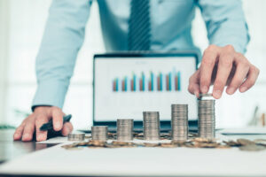 A desk with an out of focus laptop displaying a bar chart in the background. In the foreground and focused, there are 6 stacks of coins, lowest on the left and highest on the right. A man reaches over the laptop and places 2 more coins on the highest stack of coins. Clearly, when faced with the question of whether to use the cash or accrual accounting method, this business has picked the cash method.
