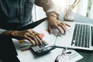 A table with a laptop on the right, a calculator to the left, and some papers scattered underneath both. One paper is titled Financial Report. A man who seems to understand basic accounting concepts is punching numbers into the calculator with his right hand and moving the touchpad with his left hand.