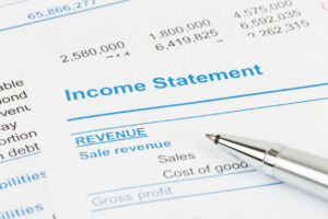 Two pieces of paper that are a business's financial reports lay on top of each other. The one on top says Income Statement and partially obscures the other. A pen juts out from the lower left of the photo. The paper on top says Income Statement and has lines for Revenue, Sale revenue, Sales, Cost of goods [sold], and Gross profit.