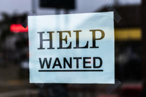 A paper sign taped to a window that says "Help Wanted," which is something a business would put out if they're looking to hire their first employee.
