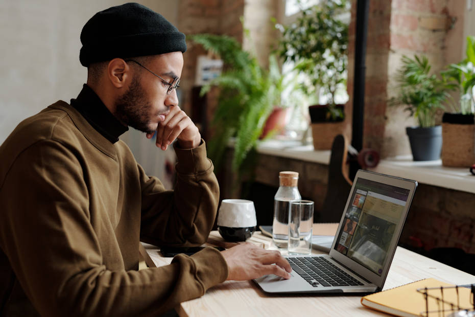 A young man in a knit cap and wearing glasses sits at a cafe looking at his laptop. His left hand is raised to his face, like he's thinking. He is moving his right index finger on the touchpad of his laptop. To his left, here's a mug likely filled with coffee and a glass bottle of water as well as an empty drinking glass. The young man is the type of worker that might make a business work through the contractor vs. employee analysis, to make sure he is being paid correctly.