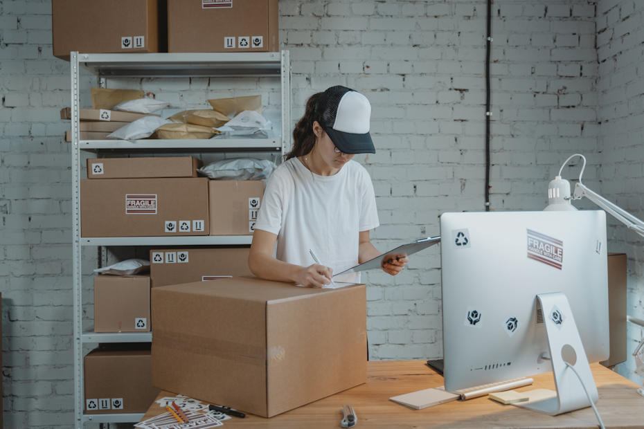 A young woman wearing a white T-shirt and a baseball cap is standing in the middle of the photo. She's looking down at a clipboard she's holding in her left hand and seems to be copying with her right hand info from the clipboard to a sticker on a cardboard box. The box is on a table, on which a large computer screen also sits. To the back of her is a shelf full of boxes. She seems to be working in a warehouse or mail room and is exactly the type of worker who is paid by the hour and for which you'll need to calculate overtime pay if she works over 40 hours a week.