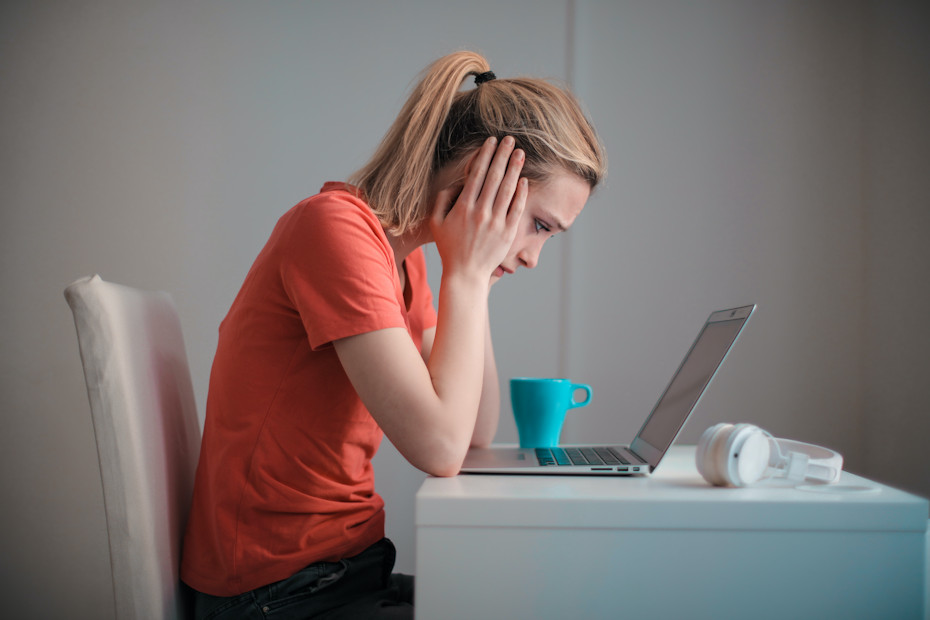 A young woman in a pony tail and wearing an orange T-shirt is sitting at a table, looking down at a laptop in front of her. Both her hands are cradling her face and she looks sad. She's just got the news that she's lost her job. Now, she'll probably need that unemployment insurance her former employer had been paying into, as a part of something every employer is required to do by law.