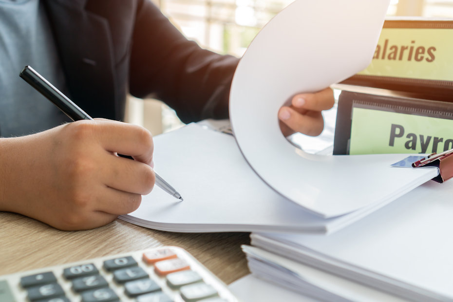 We see the hands of a person who is sitting at a table. The person is holding open a stack of paper with the left hand and writing something with the right hand. We also see the lower parts of a calculator to the photo’s bottom left. There are two large binders to the left of and slightly behind the person’s left hand. One says Salaries and the other says Payroll. Instead of doing things by hand, this person probably can save some time if they did payroll using small business payroll software.