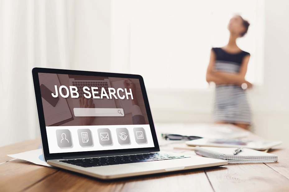 An open and powered-up laptop sits on a table along with a notebook, a pen, and some papers that are scattered behind. There's a woman standing apart from the table, with her arms crossed and looking up, but she's out of focus. The laptop is opened to an internet web page that says "Job Search" and is one of many job posting sites for employers available on the internet.