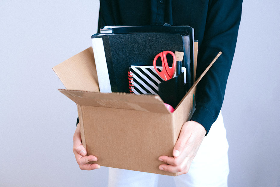 A woman in a black shirt is standing and holding with both hands a small cardboard box. In the box are some folders, a spiral notebook, and a pen holder with a pair of scissors with red handles sticking out. This is the classic photo of what happens after you let an employee go.