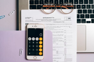 A smartphone showing a calculator app sits on top of a hard copy of Schedule 1040-SE which, in turn, is laid atop of a laptop. A pair of glasses rests half on the tax form and half on the laptop. Everything is ready for a small business owner to learn how to calculate quarterly estimated taxes.
