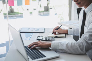 A young man in a white shirt and a black tie sits at a desk, his right hand holding a pen to jot down numbers while his left hand types info into a tax software for small businesses. A calculator and two stacks of printouts of financial reports also lay on the desk.