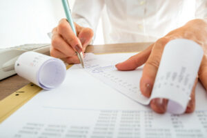 A closeup shot of a woman's hands holding a long printout of a receipt. She has circled a number on the receipt showing her business is collecting sales tax as is required. Also on the desk are a calculator, another long printout of a sales receipt, and a printout of a sales report.